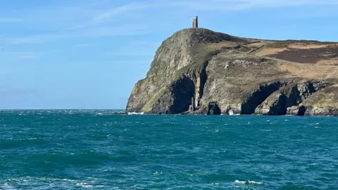 Milner's Tower on Bradda Head in Port Erin. The sea around the headland is a deep turquoise and the sky is light blue. 