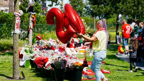PA Media A woman in a yellow top and jeans, with her hair in long purple braids, places balloons in the shape of the number 20 on a grassy area blanketed in flowers, football shirts, scarves and balloons