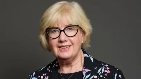 UK Parliament A woman with blonde hair and glasses in a close-up shot. She is looking at the camera and is visible from the shoulders up. She is wearing a dark jacket with a floral pattern. She is in front of a grey background.
