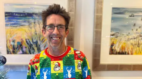 A man with spiky hair and glasses, wearing a colourful Christmas top, smiles at the camera