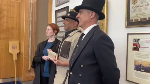 A man in a bowler hat holding a long certificate flanked by a woman with short red hair wearing a court robe and a man in a top hat and tails.