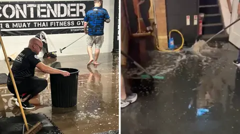 Contender Gym Two members of gym staff sweeping up water and trying to get it into a large black bucket to remove it, alongside a flooded floor.