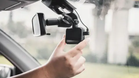 Video recording equipment in a car that has been set up on the rear-view mirror.  The photograph is a close-up of the driver's hand adjusting the kit. The view outside the car is blurred.