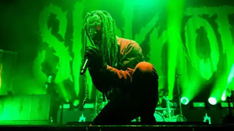 Getty Images The lead singer of Slipknot, clad in a red boilersuit, wears a gas mask-style face covering with dreadlocks attached to the top. He's at the front of a large stage, half-kneeling with a microphone clasped between both hands. Behind him, a huge Slipknot logo is bathed in eerie green light.