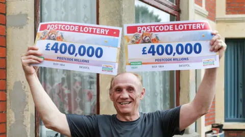 People's Postcode Lottery  Tony Labruna smiling in front of his house, holding up two postcode lottery 'cheques', both of which say £400,000. 