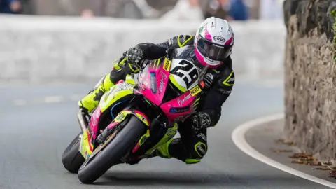 SOUTHERN 100 Naughton rounding a bend with a stone wall on the left on the Billown Course. His leathers are black and yellow, his helmet is white and he is on a pink and yellow bike.