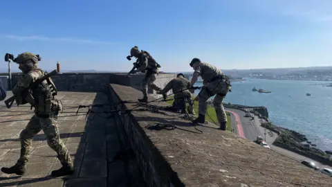 Four soldiers are seen on the wall of the Royal Citadel that overlooks Plymouth Sound. The soldiers are in military uniform and have ropes around them as they prepare to abseil down the wall. It's a sunny day with clear skies.