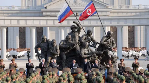 Wide shot showing North Korean leader Kim Jong Un speaking to soldiers wearing military uniforms as he stands on a monument in Pyongyang for Russian soldiers who died in the Ukraine war