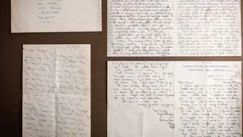 Bearnes Hampton & Littlewood The image shows an enveloped addressed to Reggie Kray in prison on the Isle of Wight, with a letter beneath. On the right there is a photo of a letter Reggie write to his mother Violet. It is on Brixton Prison headed paper and bears his prison number 058111. The front of a letter Reggie Kray wrote to his twin Ronnie from Parkhurst Prison on the Isle of Wight is also photographed. It has a date stamp 3 July 1982.