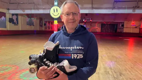 Tony has grey hair and wears thin glasses and a navy blue hoodie. He holds a pair of roller-skates and stands in the middle of a wooden rink.