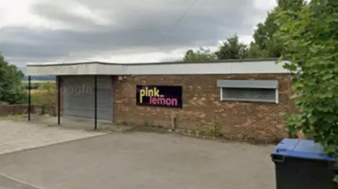 A street view of a brick, one-storey building with a white overhanging roof. There is a black sign on the building with the words 'pink lemon' written in yellow and pink. There are parking spaces in front of the building and trees on either side.