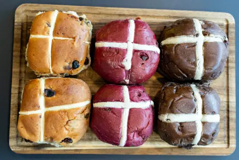 Traditional hot cross buns, red velvet and chocolate versions side by side on a wooden board.
