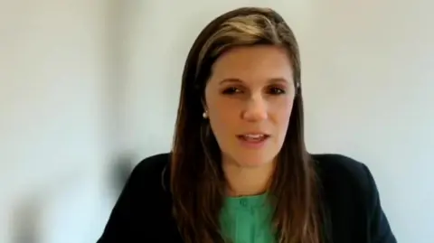 Claire Fernyhow sits facing the camera on a video call in front of a blurry background. She has long black hair and wears a green top and a black jacket.