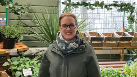 Gemma Lofthouse, a woman with brown hair and dark glasses. She is wearing a grey scarf with a light blue flowered pattern, and a dark green raincoat with a zip down the middle. Behind her is a variety of green plants, including some in black plastic pots. To the right of the image, there are a series of empty wooden boxes on a shelf, in front of a window which is covered by a white, metal security grille.