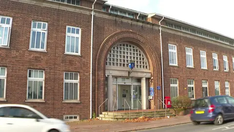 Exterior of Torquay police station. Two cars - one blue and one white - are driving along the road outside the station's front door. The building is a large red brick building with a blue police street light attached above the door.