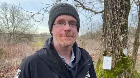 A man in a black coat and beanie hat sands in front of a tree and countryside. 