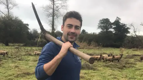 Joshua Smith is smiling at the camera as he stands in a field with nine sheep with horns behind him. He is holding a pickaxe over his shoulder that has a long wooden handle. 