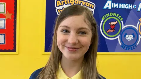 A school girl with dark blonde hair standing in a yellow school corridor.