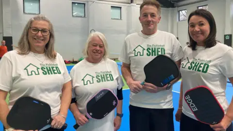 Four people stand with black pickleball rackets facing the camera, smiling. They are all wearing white t-shirts that say The Pickleball Shed. They are stood in the pickleball court which has a blue and green flooring. 
