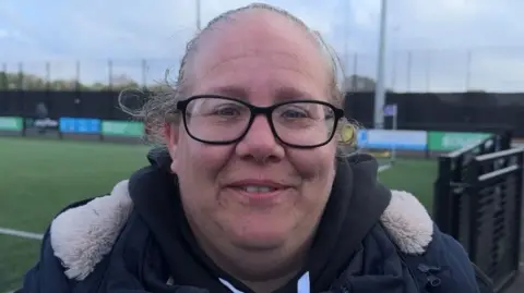 A woman with blonde hair and glasses is pictured in front of a football pitch.