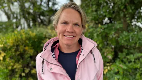 Woman with blonde hair wearing a pink jacket stands in front of green bushes