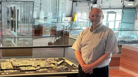 Amgueddfa Cymru A man in his late 60s on the right, stood in front of a display of treasure, including several historical gold coins from the reign of Ferdinand and Isabella of Spain, at the National Waterfront Museum in Swansea. 