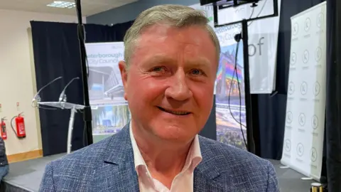 Dennis Jones is in the middle of the picture looking towards the camera smiling. He has short dark blonde hair, and is wearing a white shirt and blue suit jacket. 