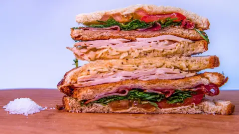 Emma Lynch/BBC The image shows a tall sandwich halved and arranged on a wooden surface against a plain, light background. Each sandwich is made with bread and filled generously with layers of sliced meat, leafy greens and tomato slices. A small pile of coarse salt sits on the wooden surface to the left of the sandwiches.