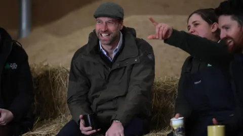The Prince of Wales, dressed in a green jacket, open-necked shirt and flat cap, sits on a hay bale while holding a mug. He is smiling as a woman to his left dressed in black with her hair tied back points something out. To the woman's left is a smiling man with dark red hair and beard.