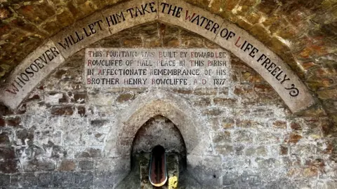 A fountain coming out of a stone wall. The wall has faded inscriptions on it.