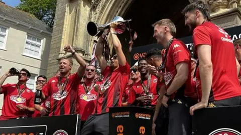 The team lift the trophy while standing on the steps of Truro Cathedral