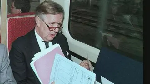 PA Media A man with papers in his hand wearing a suit and glasses is sitting on a train which has an interior from the 1970s.