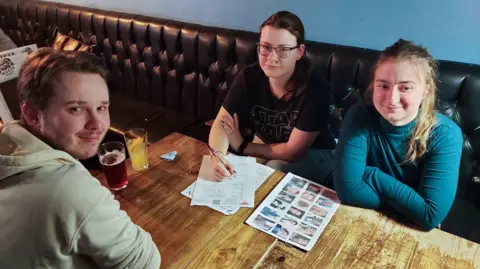 Callum with brown hair wearing a beige hoodie, Nia with long brown hair in a ponytail and black glasses wearing a Star Wars t-shirt with a pen in her hand posed to finish pub quiz questions and Anastasia with long blond hair wearing a bottle green roll neck top.