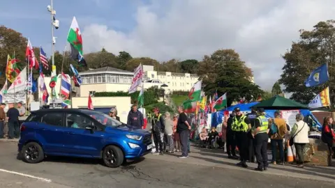 Getty Images A protest outside the hotel with flags from around the world visible as well as police officers chatting to each other. A dark blue car tries to pass.