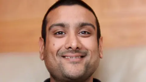 Cambridgeshire Police Neil Lakhani is smiling towards the camera. He has brown eyes, dark, cropped, hair and stubble.
