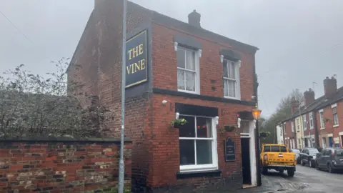 General view of The Vine Inn pub in Stoke. A red brick detached house with a black door and a side at the side that reads The Vine in gold.