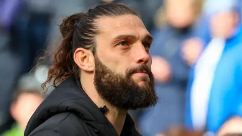 Andy Carroll has his long brown hair in a man bun. He is wearing a black hooded coat and looking into the distance. 