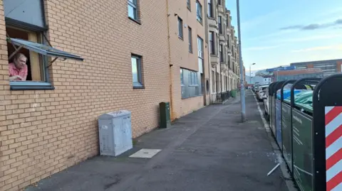Kate McNab An elderly man in a pink top sits at his open window, which is the same height and about 6ft from a bin hub. 