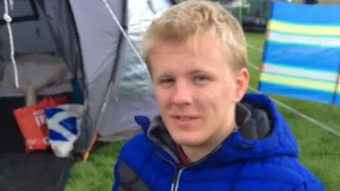 A blonde haired boy with a blue jacket smiles at the camera