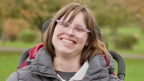 Kate is a young women in a wheelchair. She has brown shoulder length hair and wears glasses. She has on a grey puffa jacket and is smiling broadly.