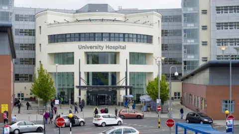UHCW (University Hospitals Coventry and Warwi)ckshire The image shows the front entrance of the University Hospital in Coventry. It is a white fronted structure set between red brick and grey clad buildings on either side, with the main hospital behind it.