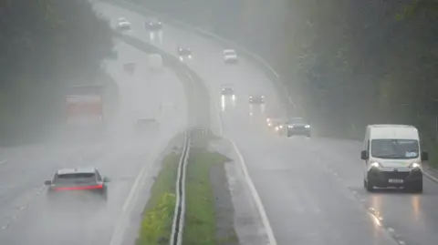 PA Media Cars, lorries and vans drive along a wet A38 in Plymouth. Most of the vehicles have their lights on. Spray from water on the dual carriageway is visible.