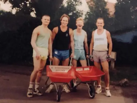 Belton Barrow Race Archive photograph of four men dressed in shorts and vests standing behind two red wheelbarrows laughing and smiling.