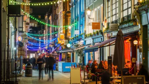 A night time view of a Soho street, showing outdoor restaurant seating and blurred pedestrians walking along the paved street. Colourful strings of green, blue and yellow lights are draped overhead between the historic shop facades.