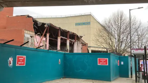 A large cream-coloured building with a small sign saying "Hollywood Cinema" is shown in the background with part-demolished buildings in the foreground with blue safety fencing in front of them.
