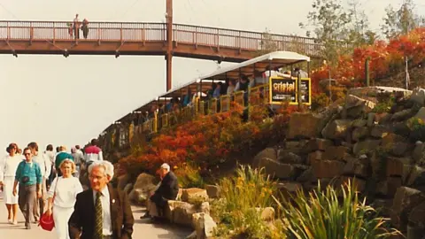People are walking along a footpath with a built-up wall of rocks on the right hand side of the image. There are people standing on a bridge which crosses the footpath. There is a train running parallel to the footpath with bushes alongside it.
