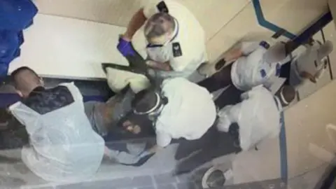 Metropolitan Police A still image from a CCTV recording of the inside of a police cell shows Nadine apparently unconscious on the floor with six officers stood around her