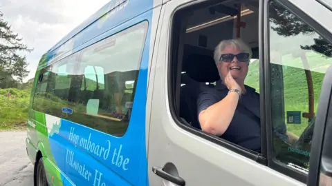 A woman sits in the driver's seat of the Ullswater Hopper. The silver minibus has blue and green branding painted in the back. The driver has short white hair and wears sunglasses and a blue polo shirt. She is smiling at the camera. The bus is parked in an area surrounded by green grass and trees.