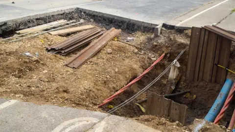 A large piece of road and pavement has been dug up and cables and pipes under the road have been exposed.