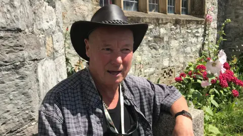 A man in a check shirt, wearing a leather stetson style hat, sitting down in front of a stone building